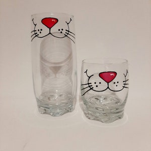 May include: Two clear glass drinking glasses with painted cat faces. The larger glass has a black outline and a red nose. The smaller glass has a black outline and a pink nose.