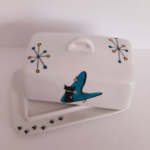 May include: White ceramic butter dish with a black cat design and blue and yellow mid-century modern accents. The lid has a handle and the base has paw prints.