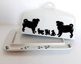 May include: White ceramic butter dish with a lid featuring black silhouettes of pugs. The lid has a handle and the base has paw prints.