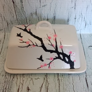 May include: White ceramic butter dish with a black and pink cherry blossom design. The lid has a handle and the dish has a base.
