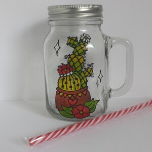 Peut inclure: Un bocal Mason en verre transparent avec un couvercle et une poignée argentés, décoré d'un motif de cactus peint à la main en jaune, vert et rouge. Une paille rayée rouge et blanche repose devant le bocal. Le bocal mesure environ 13 cm de haut.