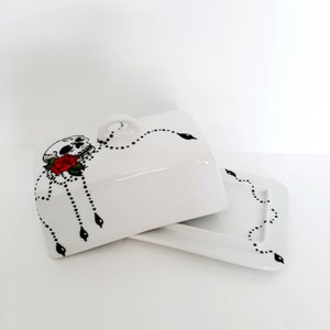 Peut inclure: Un beurrier en céramique blanche avec un motif de crâne noir et blanc et une rose rouge. Le couvercle a une poignée et la base est un plat rectangulaire.