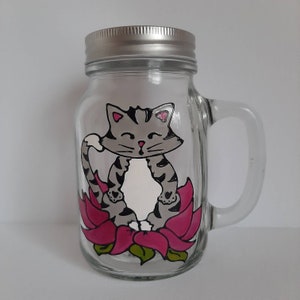 Peut inclure: Un bocal Mason en verre transparent avec un couvercle et une poignée argentés. Le bocal présente un dessin peint à la main d'un chat tigré gris en position de yoga, assis sur une fleur de lotus rose. Le chat a des rayures noires et des accents roses.