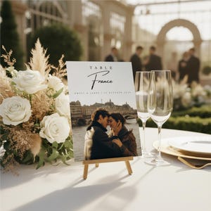 Könnte beinhalten: Eine Tischkarte mit dem Wort "Table" und "France" in Schreibschrift. Die Karte zeigt ein Foto eines sich küssenden Paares vor einem Pariser Wahrzeichen. Sie steht auf einer kleinen Holzstaffelei, mit Sektgläsern und einem Blumengesteck im Hintergrund.