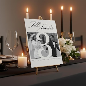 Puede incluir: Un letrero con el número de mesa de boda sobre un caballete dorado. El letrero presenta una foto en blanco y negro de una pareja, el número 3 y las palabras "Table Number" en escritura elegante. Velas y flores rodean el letrero.