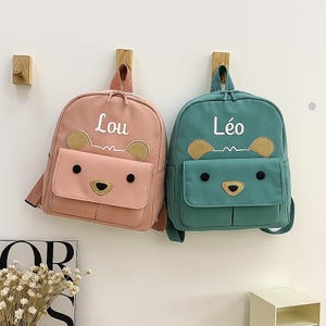 Puede incluir: Dos mochilas pequeñas con temática de oso. Una es rosa con el nombre "Lou" en blanco, y la otra es verde azulado con el nombre "Léo" en blanco. Cada mochila tiene un bolsillo frontal y detalles de cara de oso.