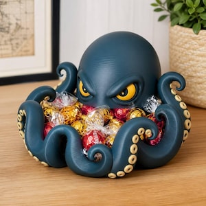 May include: A dark blue octopus-shaped bowl with a smooth, matte finish and a textured surface. The octopus has large, yellow, almond-shaped eyes and curled tentacles that form the rim of the bowl, which is filled with various wrapped candies.