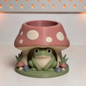 May include: A 3D printed planter shaped like a mushroom with a pink cap and white spots. A green frog sits underneath the mushroom cap on a green base. The base features small decorative elements including two tiny mushrooms and gray stones.