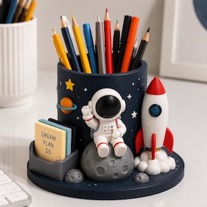 May include: A dark blue cylindrical pencil holder featuring a space theme with a 3D astronaut sitting on a gray moon, a white and red rocket ship, a yellow star, and a small orange planet. A small gray tray holds sticky notes labeled DREAM PLAN DO.