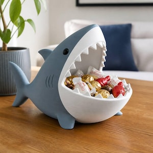 May include: A blue and white shark-shaped bowl with an open mouth filled with various wrapped sweets. The shark features a smooth matte finish, a dorsal fin, and a tail, with a white interior mouth area containing several gold and red foil-wrapped treats.