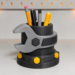 May include: A black cylindrical pencil holder featuring a silver wrench design wrapped around the center. The base is decorated with yellow hexagonal bolt heads. Inside the holder are four writing instruments: a yellow pencil, a black pen, a gray marker, and a yellow highlighter.