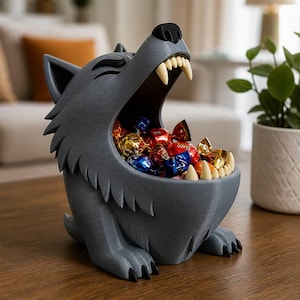 May include: A gray wolf-shaped bowl with a wide-open mouth featuring sharp, off-white teeth. The interior of the bowl is filled with various colorful, foil-wrapped candies. The wolf has a textured surface, pointed ears, and black claws on its paws.