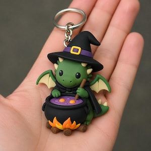 May include: A green dragon keychain featuring a black witch hat with a purple band and yellow buckle. The dragon wears a black cape and stirs a black cauldron filled with purple liquid and yellow bubbles, set over a small orange and yellow fire.