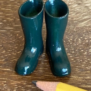 May include: A pair of miniature dark green rubber boots. The boots are glossy and have a classic wellington boot shape. A yellow pencil is in the foreground, providing a size reference. The boots are likely for a doll or miniature display.