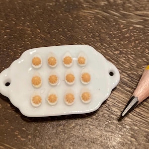 May include: Miniature white ceramic serving platter with 12 small, egg-shaped appetizers topped with orange-colored filling. A yellow pencil with a black tip is in the background. The platter has two small holes on either side.