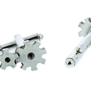 May include: A pair of silver cufflinks featuring a gear design. The cufflinks are made of metal and have a polished finish.