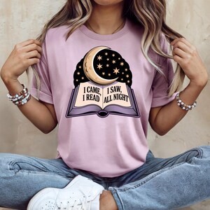 May include: A light purple t-shirt featuring a graphic of an open book with a crescent moon and stars. The text on the book reads "I CAME, I SAW, I READ ALL NIGHT". The shirt has short sleeves and a relaxed fit.