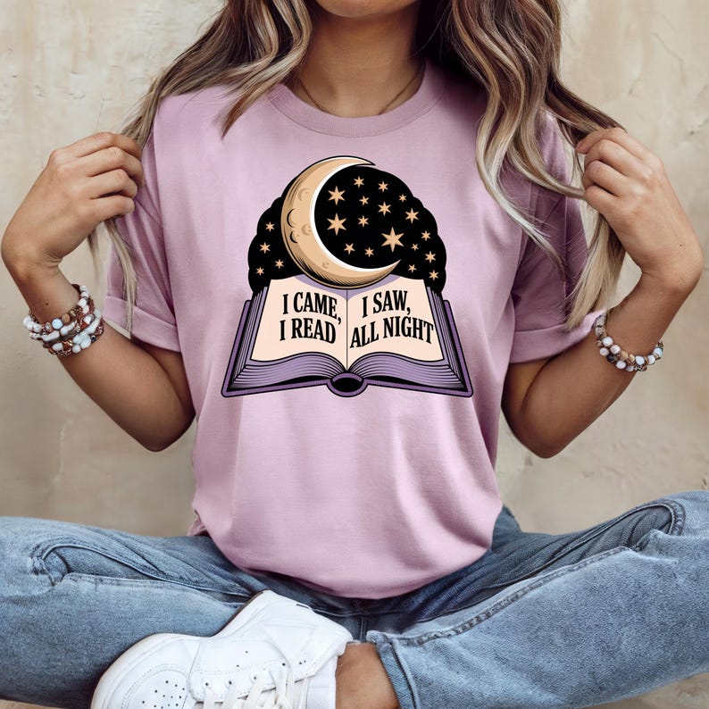 May include: A light purple t-shirt featuring a graphic of an open book with a crescent moon and stars. The text on the book reads "I CAME, I SAW, I READ ALL NIGHT". The shirt has short sleeves and a relaxed fit.