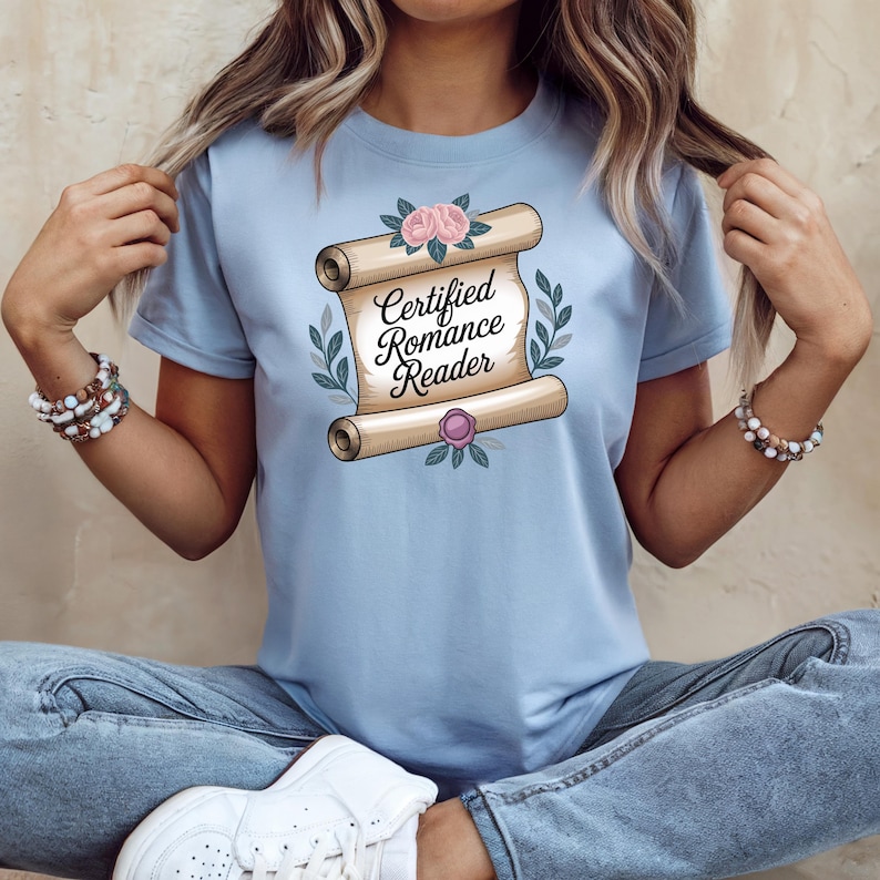May include: Light blue t-shirt featuring a scroll design with the text "Certified Romance Reader". The scroll is decorated with pink roses, green leaves, and a purple seal. The shirt is made of a soft material.
