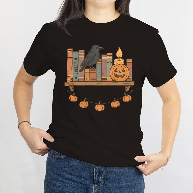 May include: Black t-shirt featuring a Halloween-themed graphic. The design includes a shelf with books, a crow, a jack-o'-lantern candle, and a string of pumpkin lights. The word "SPELLS" is visible on one of the book spines.