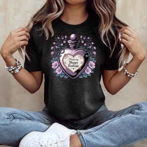 May include: Dark gray t-shirt featuring a heart-shaped potion bottle design. The bottle is purple and black, surrounded by flowers and vines. The text "Healed by Happy Endings" is inside the bottle.