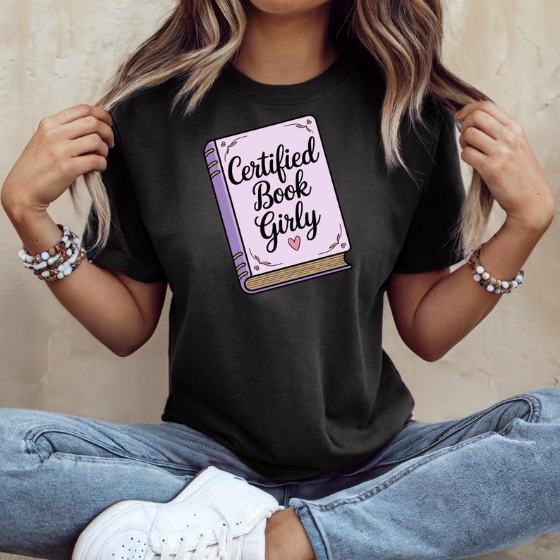 May include: Black t-shirt featuring a graphic of a book with the text "Certified Book Girly" in a script font. The book graphic is light purple with gold-colored edges and a pink heart. The shirt is a casual style.
