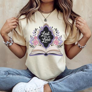 May include: Cream-colored t-shirt featuring the text "Lost to the Lore" in a decorative frame above an open book. The design includes pink roses, purple accents, and black leaves. The shirt is a casual wear item.