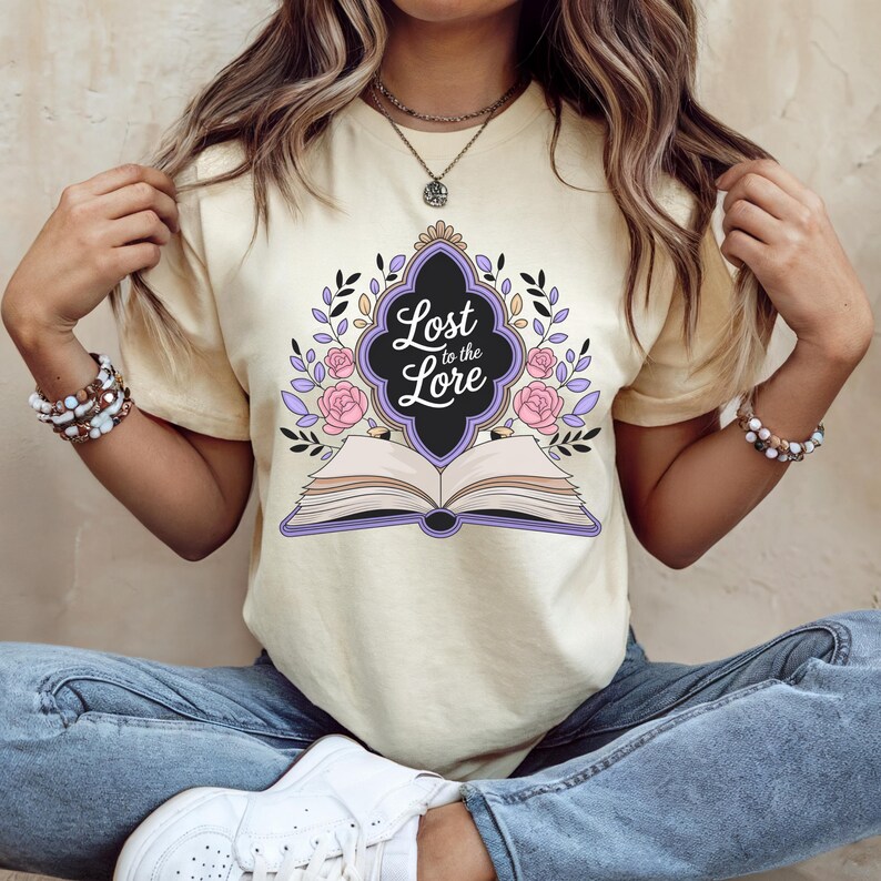 May include: Cream-colored t-shirt featuring the text "Lost to the Lore" in a decorative frame above an open book. The design includes pink roses, purple accents, and black leaves. The shirt is a casual wear item.