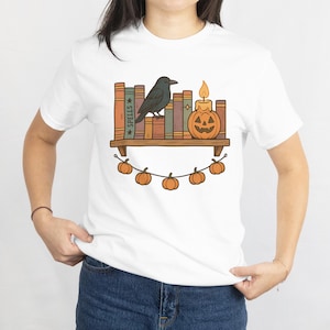 May include: White t-shirt featuring a Halloween-themed graphic. The design includes a bookshelf with books, a black crow, a jack-o'-lantern candle, and a string of pumpkins. The word "SPELLS" is visible on one of the book spines.
