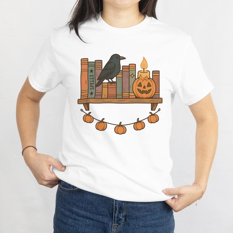 May include: White t-shirt featuring a Halloween-themed graphic. The design includes a bookshelf with books, a black crow, a jack-o'-lantern candle, and a string of pumpkins. The word "SPELLS" is visible on one of the book spines.