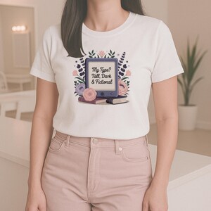 May include: White t-shirt with a design featuring an e-reader, books, and floral elements. The text on the e-reader reads "My Type? Tall, Dark & Fictional". The design incorporates pink, purple, and blue, creating a literary theme.