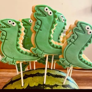 May include: Green dinosaur-shaped biscuits on sticks, arranged on a watermelon half. The biscuits have white eyes and are decorated with green icing. A fun treat for a party or celebration.
