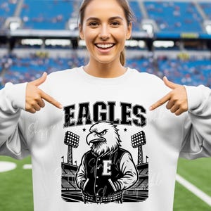 May include: White sweatshirt with a black and white graphic of an eagle wearing a letterman jacket. The word "EAGLES" is above the eagle. The background shows a stadium with stadium lights.