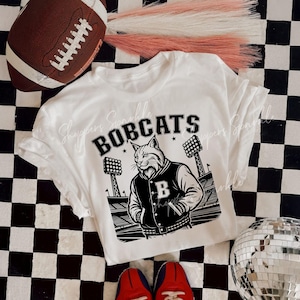 May include: White t-shirt with a black and white graphic of a bobcat wearing a letterman jacket. The word "BOBCATS" is printed above the graphic. A football and red shoes are also in the image.