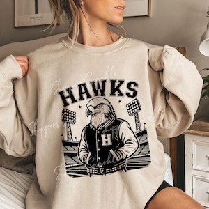 May include: Beige sweatshirt with the word "HAWKS" above a graphic of an eagle wearing a letterman jacket with the letter "H". The eagle is standing in front of a stadium with stadium lights. The sweatshirt is a neutral color.