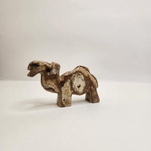 Op de afbeelding: Een kleine, gebeeldhouwde kameelfiguur. De sculptuur is gemaakt van een steenachtig materiaal met een mix van bruine, beige en witte kleuren. De kameel staat met zijn hoofd omhoog en heeft een gedetailleerde textuur. De figuur is een decoratief item.