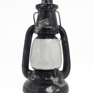 May include: A decorative lantern with a black and white marbled design. The lantern features a pointed top, a central white section, and a base with a circular design. The handle is made of a dark metal.