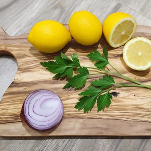 Puede incluir: Una tabla de cortar de madera con asa muestra tres limones amarillos enteros y dos mitades de limón. Perejil verde fresco y una cebolla roja en rodajas también están en la tabla. La tabla tiene una veta de madera natural.