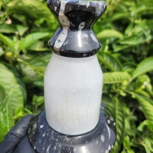 May include: A decorative bottle stopper with a black and white patterned top and base, and a white body. The bottle stopper is made of stone and has a unique shape. The background is blurred green foliage.