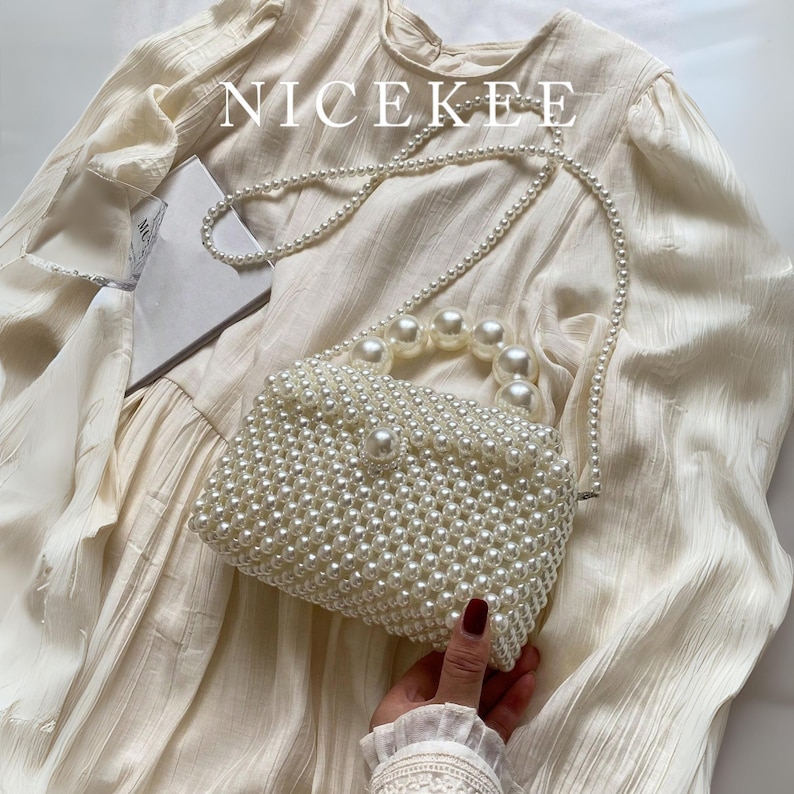 May include: A white beaded handbag with a pearl handle and strap. The bag is rectangular and covered in small, round beads. The word "NICEKEE" is printed at the top. The bag is resting on a cream-colored dress.