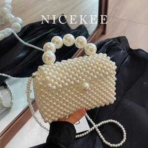 May include: A rectangular, off-white beaded handbag with a pearl handle and a long pearl strap. The bag is covered in small, round beads and features a large pearl clasp. The word "NICEKEE" is printed in white at the top.
