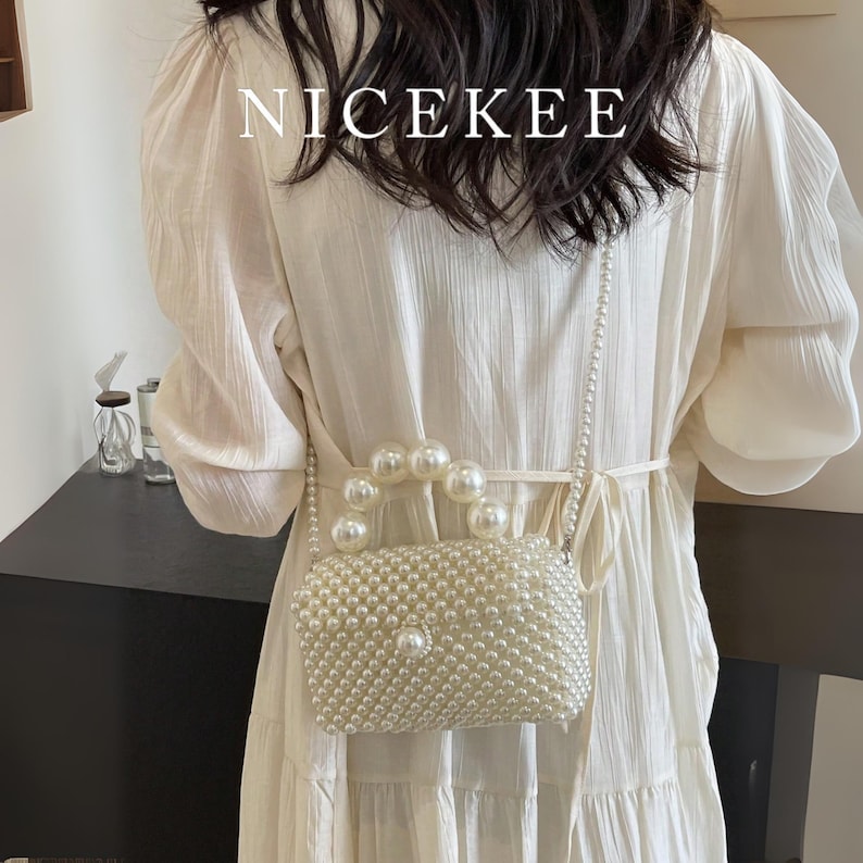 May include: A white pearl beaded handbag with a pearl handle and a pearl chain strap. The bag is rectangular with a single pearl accent on the front. The word "NICEKEE" is printed in white at the top.