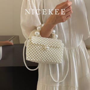 May include: A white beaded handbag with a pearl handle and a long pearl strap. The bag is rectangular with a flap closure and is covered in small, round beads. The text "NICEKEE" is displayed in white letters at the top.