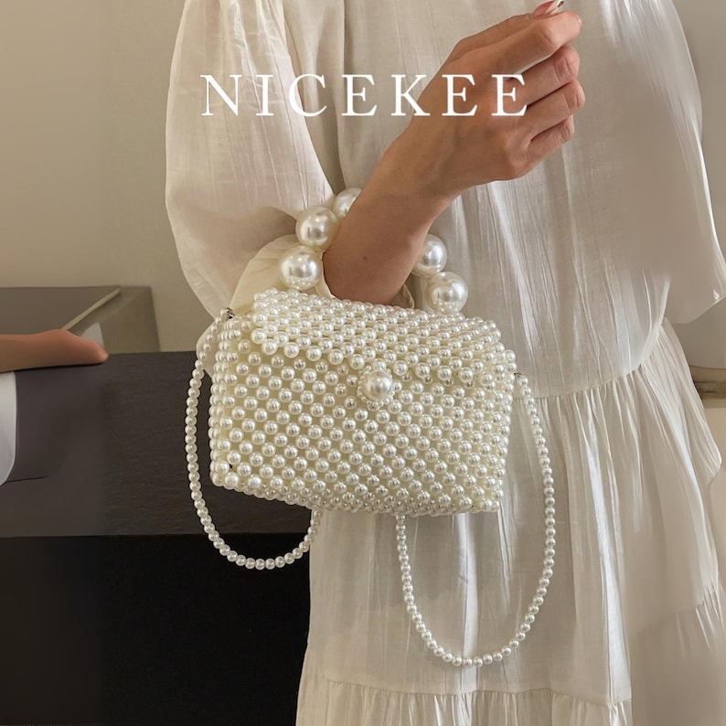 May include: A white beaded handbag with a pearl handle and a long pearl strap. The bag is rectangular with a flap closure and is covered in small, round beads. The text "NICEKEE" is displayed in white letters at the top.