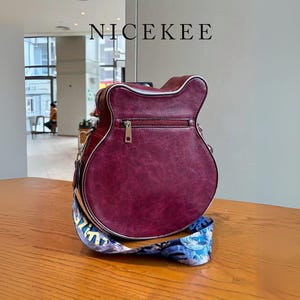 May include: A burgundy guitar-shaped handbag with a silver zipper and trim. The bag has a colorful patterned strap and the word "NICEKEE" is printed at the top. The bag is sitting on a wooden table.