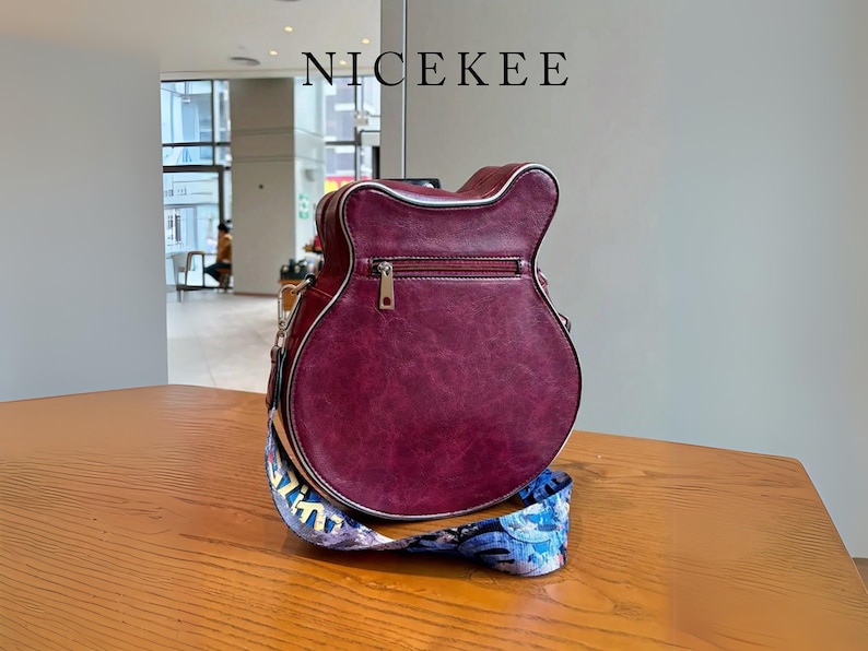 May include: A burgundy guitar-shaped handbag with a silver zipper and trim. The bag has a colorful patterned strap and the word "NICEKEE" is printed at the top. The bag is sitting on a wooden table.
