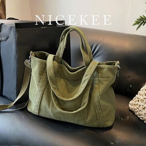 Peut inclure: Sac fourre-tout en velours côtelé vert olive avec bandoulière et poignées supérieures. Le sac a une poche avant et le mot "NICEKEE" est imprimé en haut. Le sac est posé sur une surface noire.