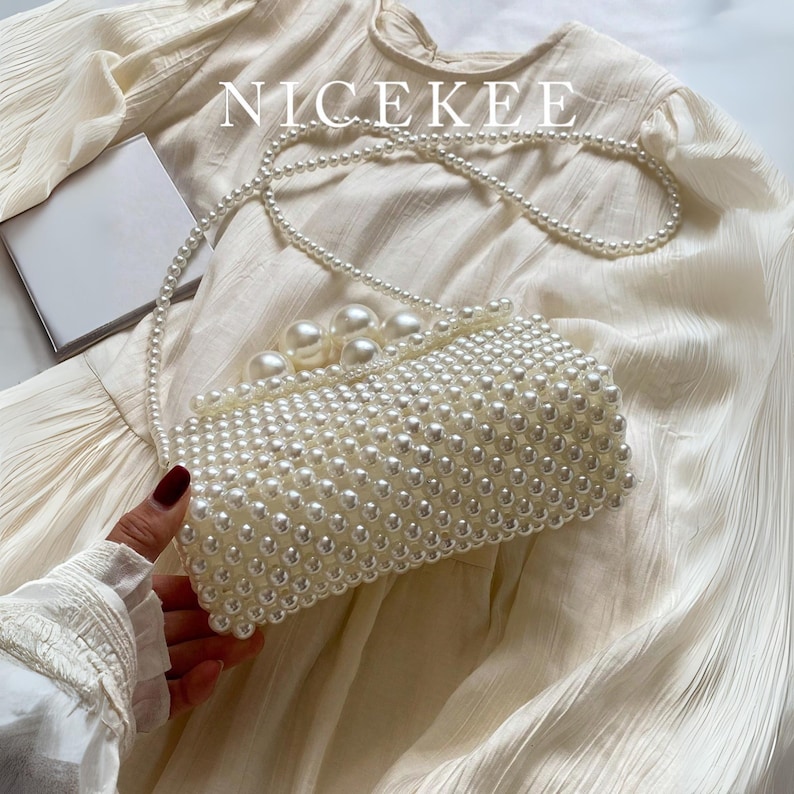 May include: A rectangular, off-white clutch covered in small, round pearls. The clutch has a pearl strap and is embellished with larger pearls on the top. The word "NICEKEE" is printed in white across the top.