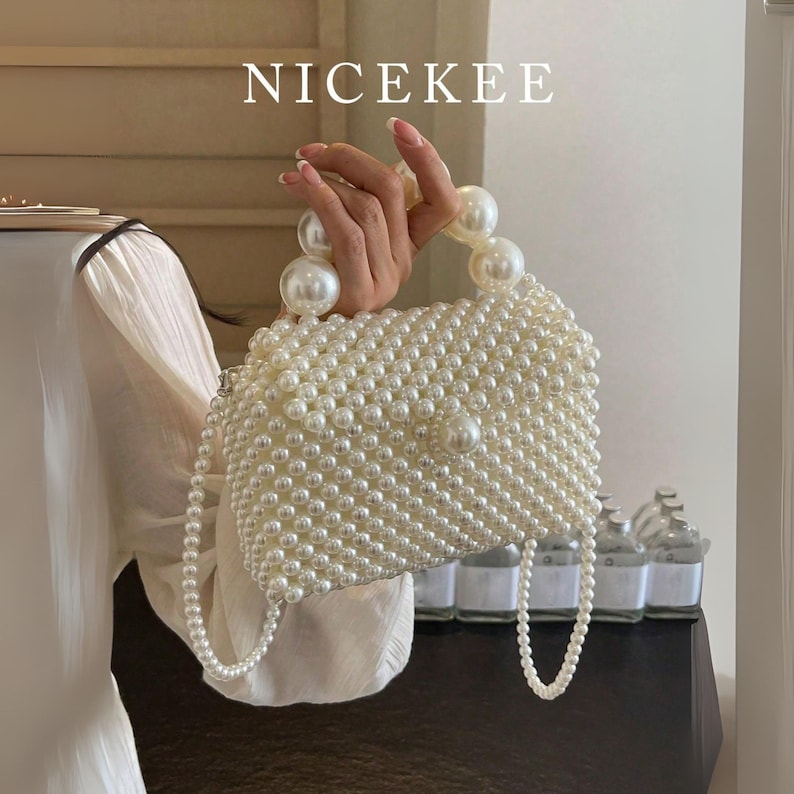 May include: A white pearl beaded handbag with a pearl handle and a pearl shoulder strap. The handbag is rectangular with a flap closure. The word "NICEKEE" is displayed at the top of the image.