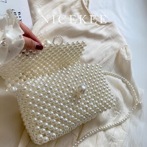 May include: A white beaded clutch bag with a pearl strap. The rectangular bag is covered in small, round, white beads. The flap is secured with a large pearl. The word "NICEKEE" is printed in white on the top of the image.